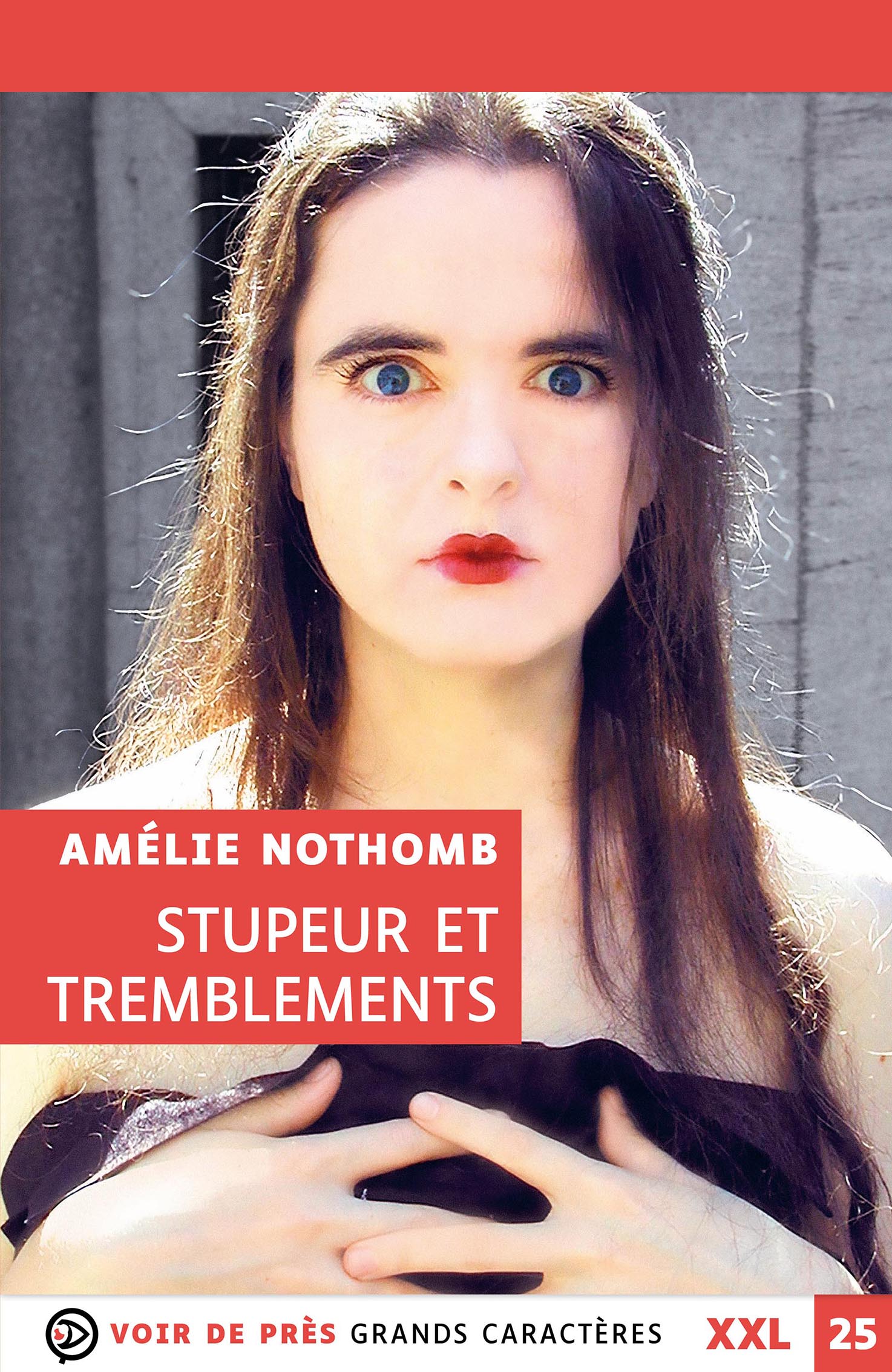 Portrait en gros plan d’une jeune femme au regard intense, aux yeux très ouverts et aux lèvres rouges, tenant ses mains près de sa poitrine, sur un fond neutre légèrement flou.